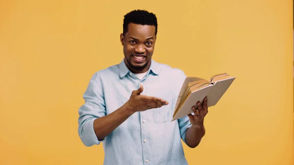 Glücklich afrikanisch-amerikanischer Mann im blauen Hemd zeigt mit der Hand auf Buch isoliert auf gelb — Stockfoto