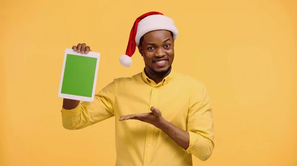 Happy african american man in santa hat pointing with hand at digital tablet with green screen isolated on yellow — Stock Photo