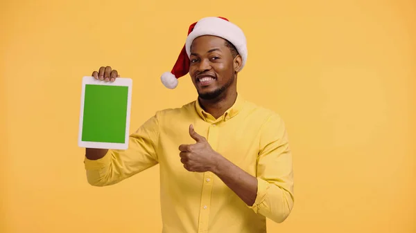 Happy african american man in santa hat showing thumb up and holding digital tablet with green screen isolated on yellow — Stock Photo