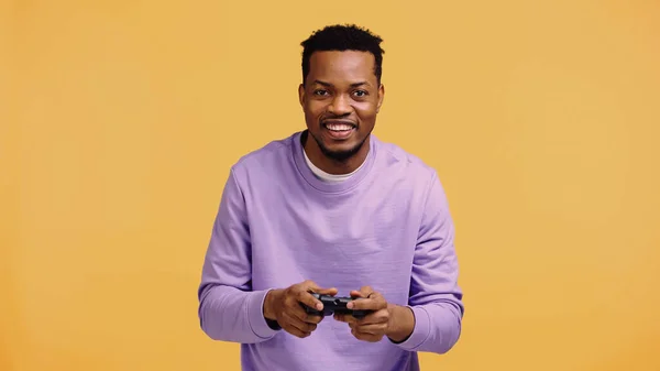 КИЕВ, УКРАИНА - 22 НОЯБРЯ 2019 года: веселый африканский американец, играющий в видеоигру изолированный на желтом — стоковое фото