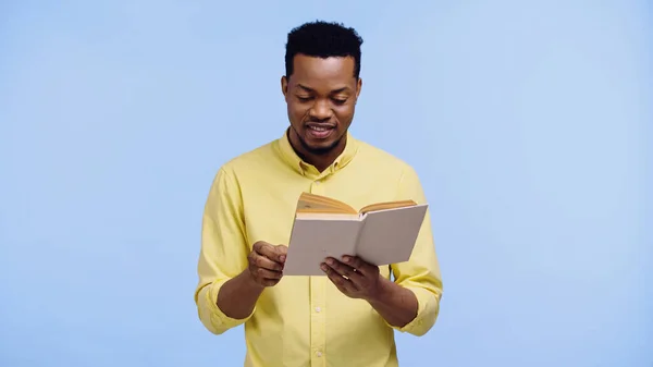 Щасливий афроамериканський чоловік у жовтій сорочці читання книги ізольовані на синьому — стокове фото