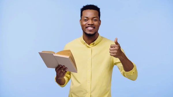 Homem americano africano feliz na camisa amarela que mostra o polegar acima e que prende o livro isolado no azul — Fotografia de Stock