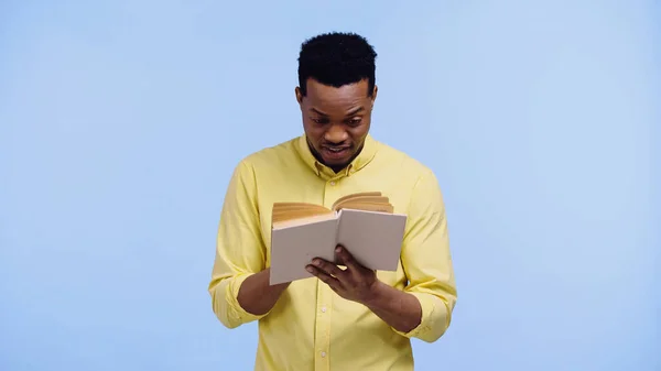 Збуджений афроамериканський чоловік у жовтій сорочці читає книгу ізольовано на синьому — стокове фото