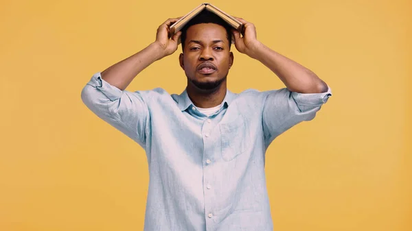 Tired african american man in blue shirt holding book on head isolated on yellow — Stock Photo