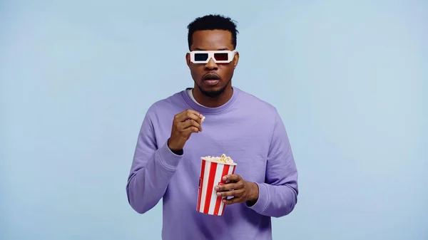 Focused african american man in 3d glasses watching movie and holding popcorn bucket isolated on blue — Stock Photo