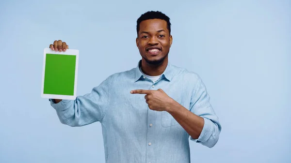 Hombre afroamericano feliz apuntando a la tableta digital con la pantalla verde aislada en azul - foto de stock