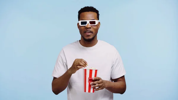 Hombre afroamericano enfocado en gafas 3d viendo películas y sosteniendo palomitas aisladas en azul - foto de stock