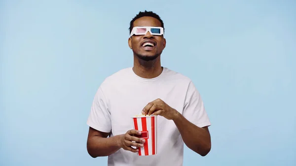 Hombre afroamericano feliz en gafas 3d viendo la película y sosteniendo palomitas de maíz cubo aislado en azul - foto de stock