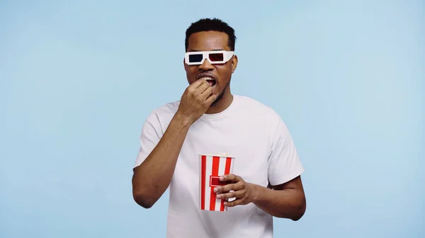 Hombre afroamericano enfocado en gafas 3d viendo películas y comiendo palomitas aisladas en azul - foto de stock