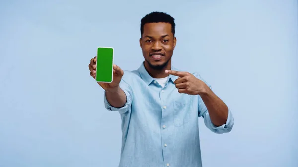 Felice uomo afroamericano in camicia che punta verso smartphone con schermo verde isolato su blu — Foto stock