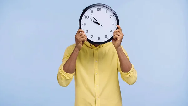 Upset african american man in yellow shirt hiding behind clock isolated on blue — Stock Photo