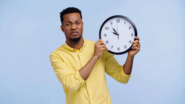 Displeased african american man in yellow shirt holding clock isolated on blue — Stock Photo