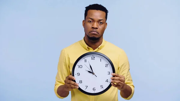 Homme afro-américain bouleversé en chemise jaune tenant horloge isolé sur bleu — Photo de stock