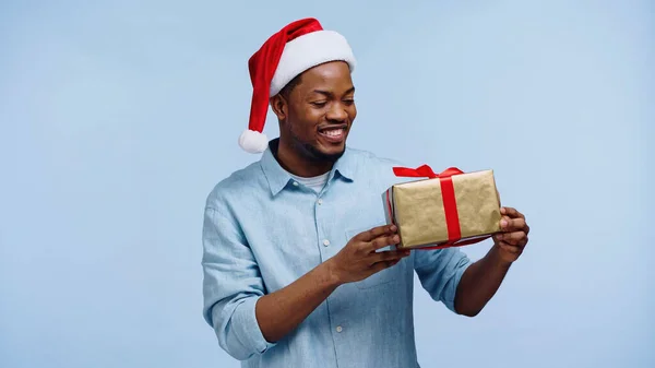 Feliz homem americano africano em santa chapéu segurando caixa de presente de Natal isolado em azul — Fotografia de Stock