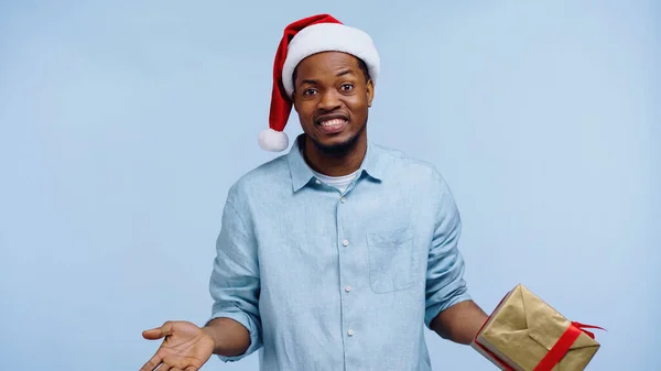 Confused african american man in santa hat holding christmas gift box and showing shrug gesture isolated on blue — Stock Photo
