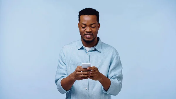 Hombre afroamericano feliz en camisa sosteniendo el teléfono móvil aislado en azul - foto de stock
