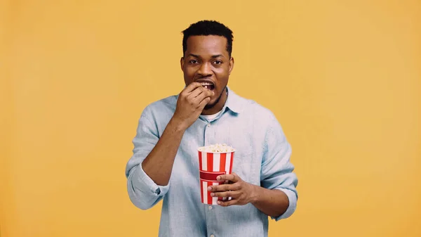 Hombre afroamericano feliz comiendo palomitas de maíz y mirando la cámara aislada en amarillo - foto de stock