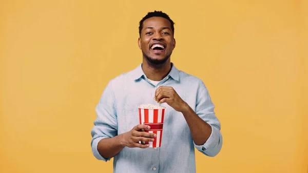 Hombre afroamericano feliz sosteniendo palomitas de maíz y riendo mientras mira la cámara aislada en amarillo - foto de stock