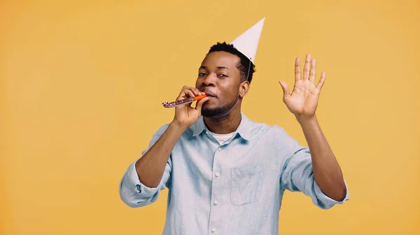Афроамериканський чоловік у святковій шапці дме партійний ріг і жестикулює ізольовано на жовтому — стокове фото