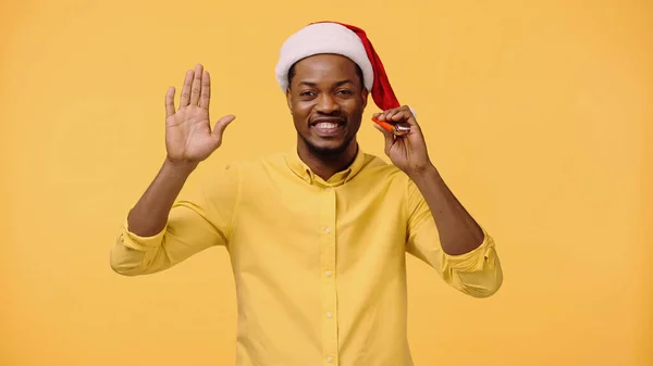 Happy african american man in santa hat waving hand isolated on yellow — Stock Photo