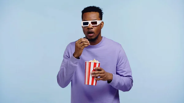 Sorprendido hombre afroamericano en gafas 3d viendo película y sosteniendo palomitas de maíz cubo aislado en azul - foto de stock