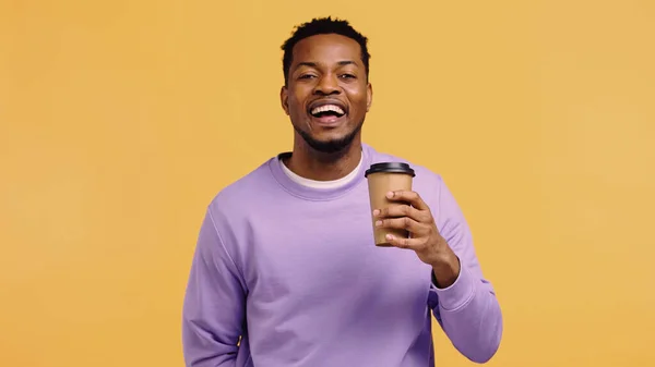 Alegre hombre afroamericano en jersey púrpura sosteniendo taza de papel con café para ir aislado en amarillo - foto de stock