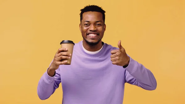 Homem americano africano alegre em pulôver roxo segurando café para ir e mostrando como isolado no amarelo — Fotografia de Stock