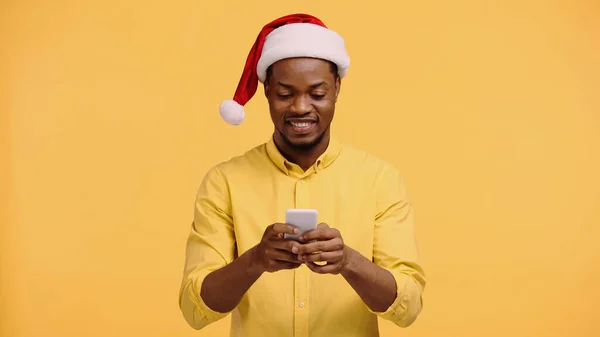 Happy african american man in santa hat using smartphone isolated on yellow — Stock Photo