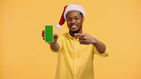 Homem americano africano feliz em santa hat apontando para smartphone com tela verde isolada em amarelo — Fotografia de Stock