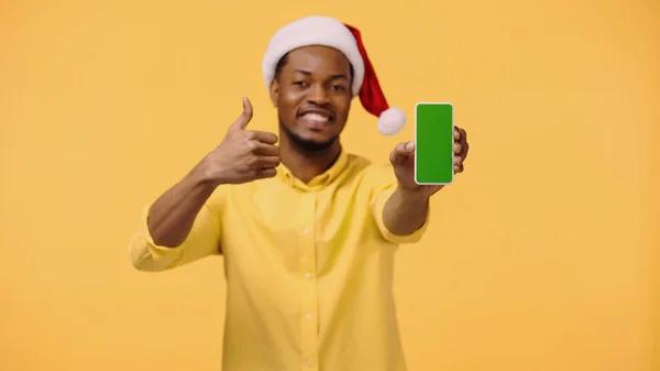 Счастливый африканский американец в шляпе Санты держит смартфон с зеленым экраном, показывая, как изолированные на желтый — стоковое фото
