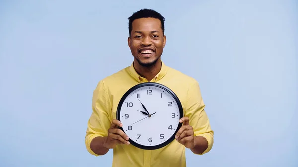 Homme afro-américain joyeux tenant horloge et regardant la caméra isolée sur bleu — Photo de stock