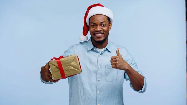 Felice uomo afroamericano in cappello da Babbo Natale avvolto presente mentre mostra come isolato sul blu — Foto stock