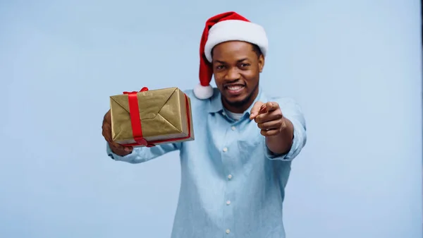 Glücklicher afrikanisch-amerikanischer Mann mit Weihnachtsmütze und umwickeltem Geschenk, während er mit dem Finger auf die Kamera zeigt, isoliert auf blauem Grund — Stockfoto