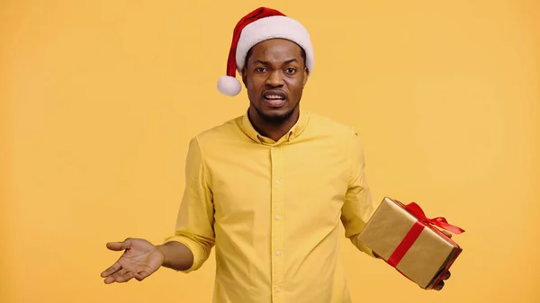 Confused african american man in santa hat holding present and showing shrug gesture isolated on yellow — Stock Photo