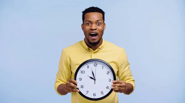 Amazed african american man holding clock and looking at camera isolated on blue — Stock Photo