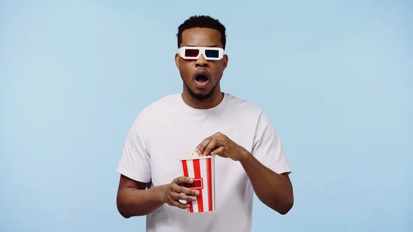 Sorprendido africano americano hombre en 3d gafas viendo película y sosteniendo palomitas cubo aislado en azul - foto de stock