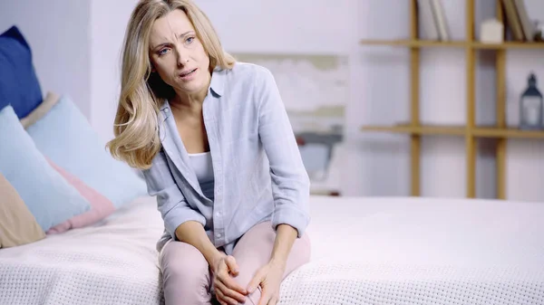 Donna bionda che soffre di dolore al ginocchio mentre è seduta a letto a casa — Foto stock
