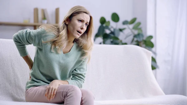 Worried woman having pain in back while sitting on couch in modern living room — Stock Photo