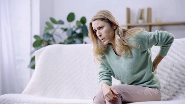 Blonde Frau hat Rückenschmerzen, während sie auf der Couch im modernen Wohnzimmer sitzt — Stockfoto