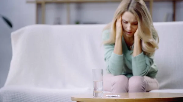 Bicchiere d'acqua e pillole sul tavolino vicino alla donna con mal di testa su sfondo sfocato — Foto stock