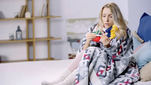 Sick woman sitting on bed under colorful blanket and measuring temperature with thermometer — Stock Photo