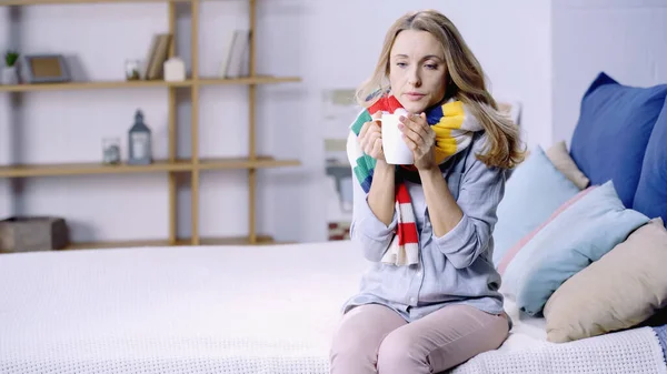 Sick and upset woman in colorful scarf holding cup of tea and sitting on bed at home — Stock Photo