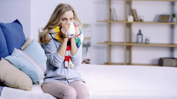 Больная женщина в красочном шарфе пьет чай и сидит на кровати дома — стоковое фото