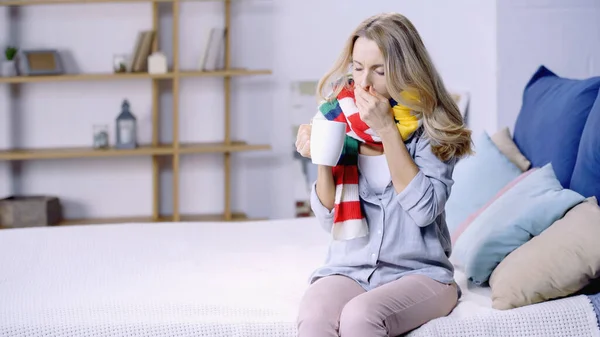 Sick woman in colorful scarf drinking tea and coughing while sitting on bed at home — Stock Photo