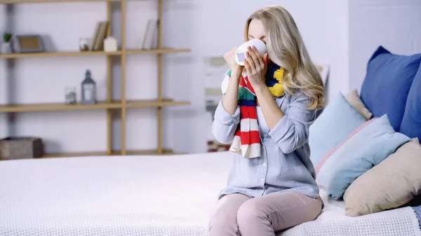 Diseased woman in colorful scarf drinking tea and sitting on bed at home — Stock Photo