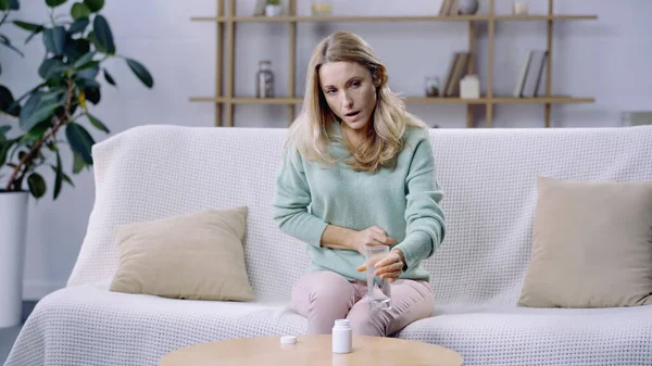 Mujer agotada que sufre de dolor abdominal y sosteniendo un vaso de agua cerca de la botella con pastillas — Stock Photo