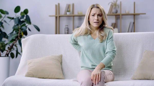 Blonde woman having pain in back while sitting on sofa in living room — Stock Photo