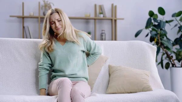 Blonde woman suffering from pain and putting pillow behind back while sitting on sofa — Stock Photo