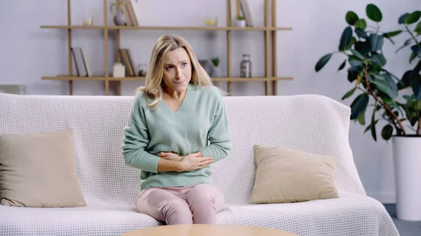 Exhausted woman having cramps and suffering from abdominal pain while sitting on couch — Stock Photo