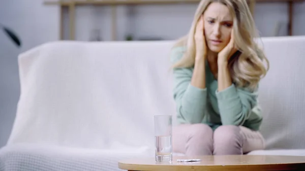 Glas Wasser und Pillen auf Couchtisch in der Nähe von Frau mit Migräne auf verschwommenem Hintergrund — Stockfoto